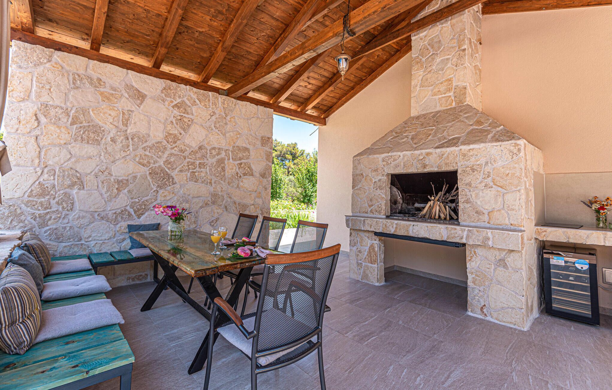 An outdoor patio with stone walls, a wooden ceiling, a stone fireplace, and a dining table with chairs.