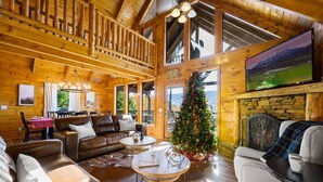 A cozy log cabin interior with a fireplace, a Christmas tree, leather sofas, and large windows offering scenic views. - 5 Decks | Hot Tub | Views | Fire Pit | Pool Table (Sevierville)