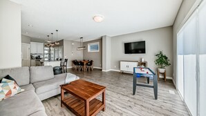 A modern living room with a sofa, coffee table, dining area, kitchen, and a foosball table. - Spacious Home w/ Sauna & Fenced Yard w/ BBQ, Pet-Friendly, Sleeps 10, King Beds (Calgary)