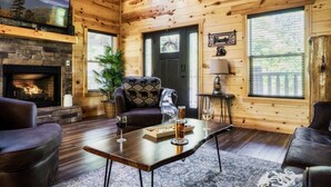 Acogedor interior de cabaña de madera con chimenea de piedra, sofás de cuero, una mesa de comedor con vasos y un ventanal con vistas al exterior.