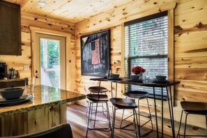 A wooden cabin interior with a kitchen area, bar stools, and a large window with blinds.
