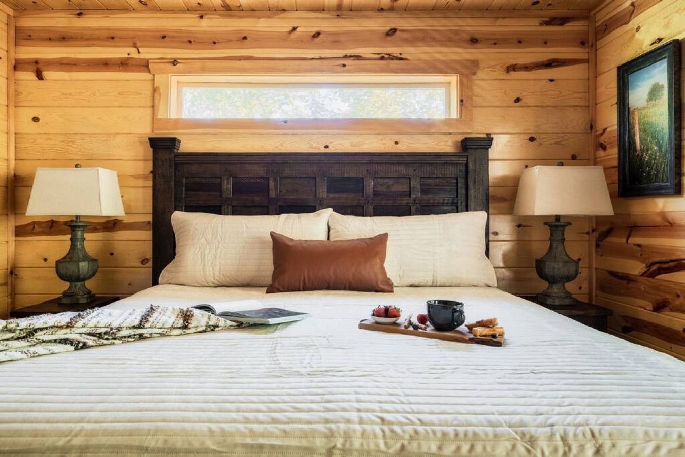 A wooden cabin bedroom with a large bed, two bedside lamps, a window, and a painting on the wall.