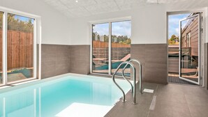 Una piscina interior moderna con paredes de vidrio transparente, una escalera metálica y vistas a una terraza de madera y árboles al exterior.