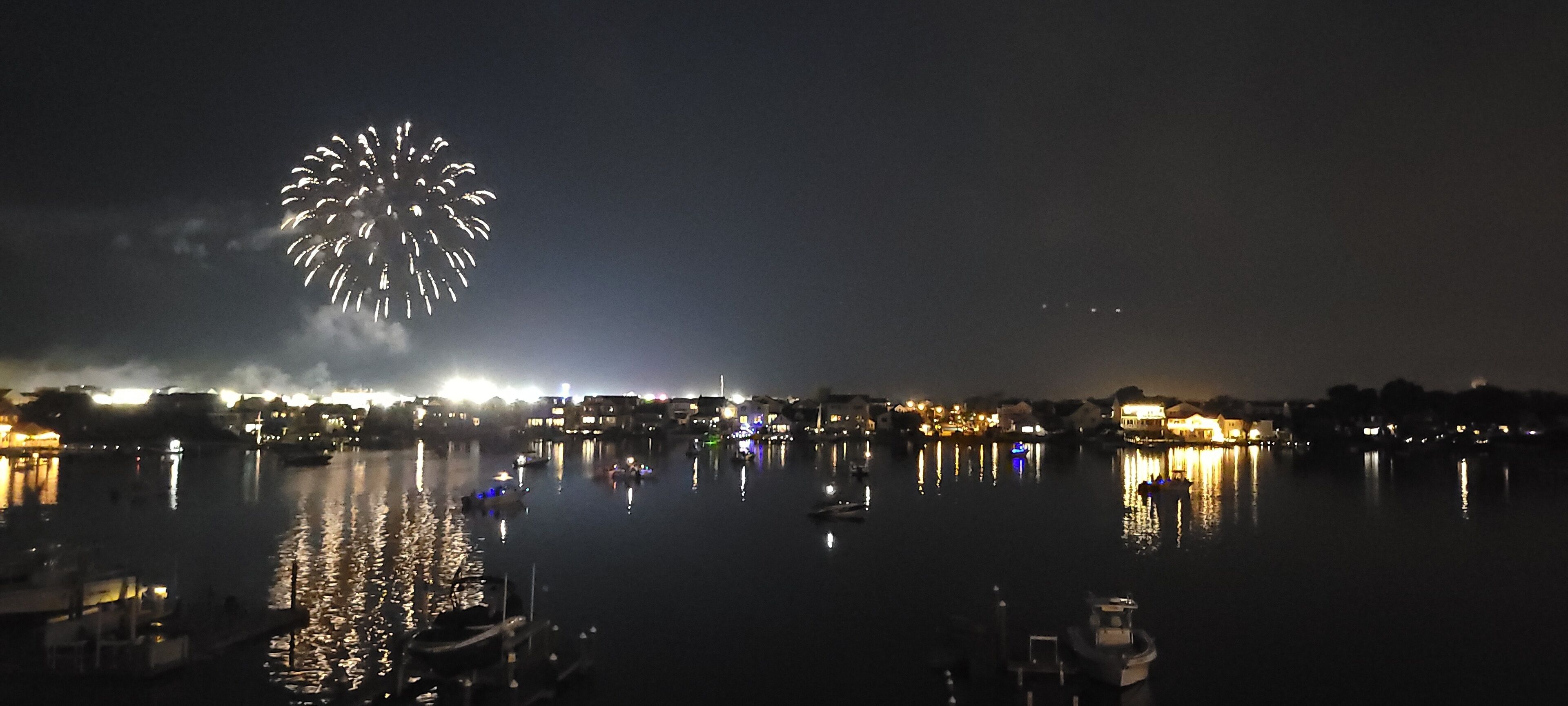 Une vue nocturne d’un front de mer, avec des bateaux et des bâtiments dont les reflets se mirent dans l’eau.