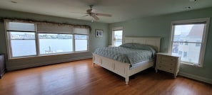 A bedroom with a bed, nightstands, a ceiling fan, and large windows overlooking a body of water.