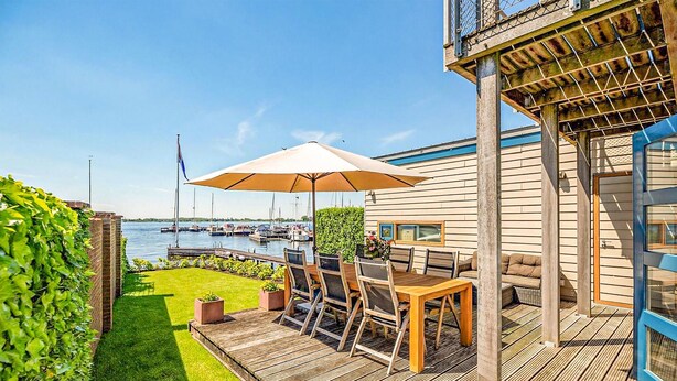 A wooden deck with outdoor furniture, a view of the marina, and a building with a blue accent wall. - Nice home in Breukeleveen with WiFi (Breukeleveen)