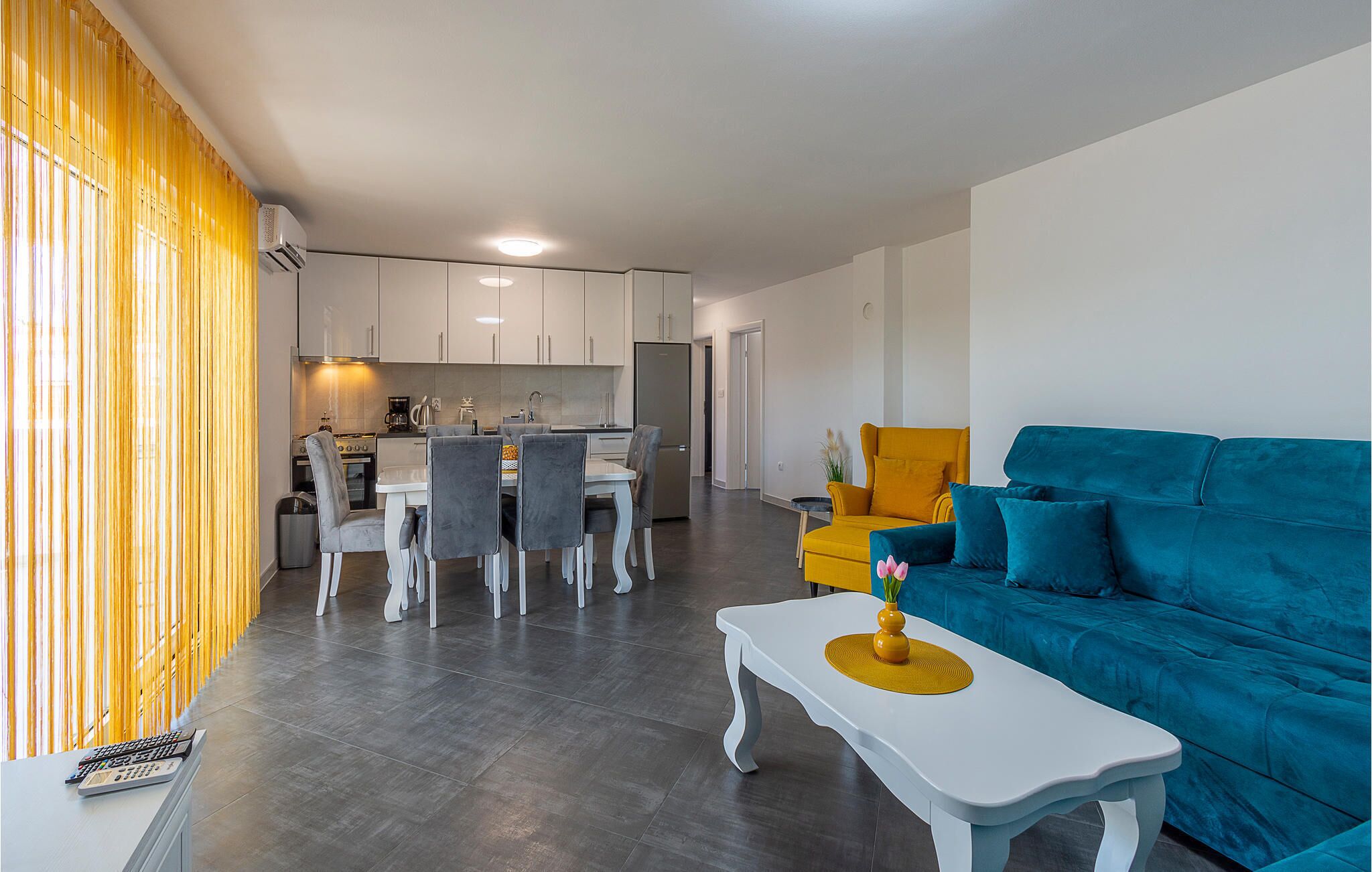 Ein modernes Wohnzimmer mit einem blauen Sofa, einem weißen Couchtisch und einem Essbereich mit weißem Tisch und Stühlen.
