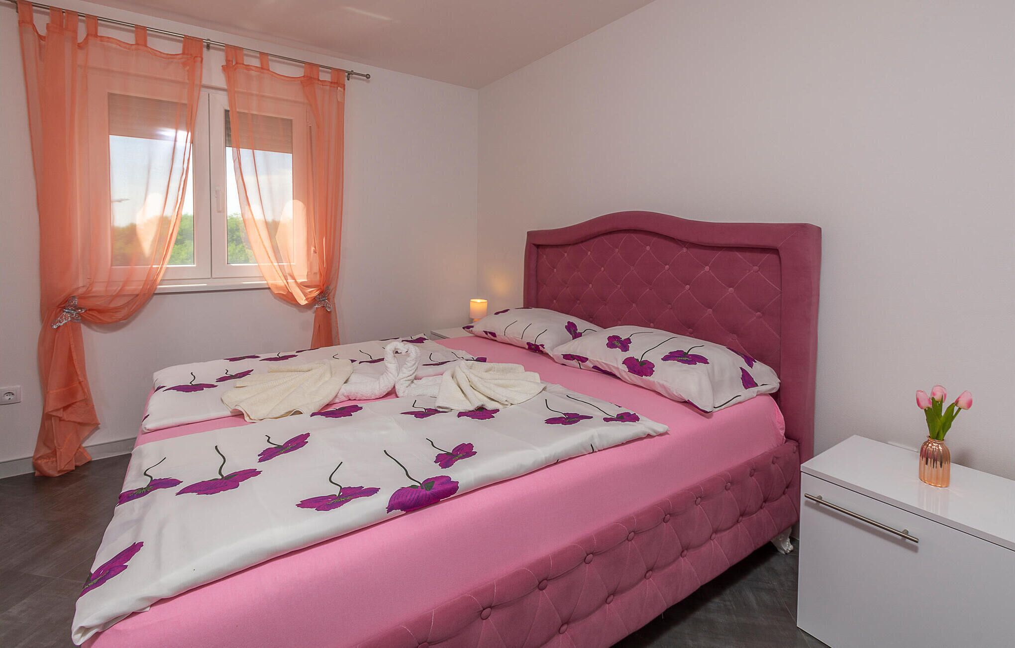 A bedroom with a pink bed, white pillows with purple floral patterns, and a white nightstand with a vase of flowers.