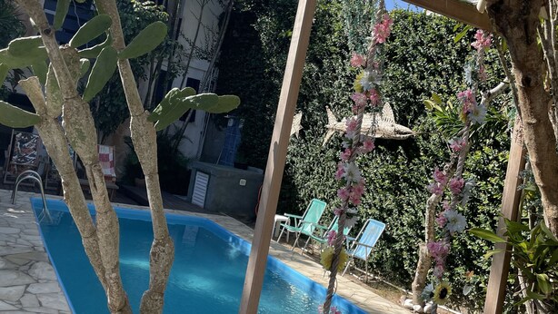 A pool area with a wooden swing chair, a small wooden structure, and a view of a building in the background. - Pousada Bicho do Mar (Arraial do Cabo)