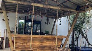 A rustic outdoor seating area with a wooden bench and tables, a metal roof, and a backdrop of trees and a building. - Pousada Bicho do Mar (Arraial do Cabo)