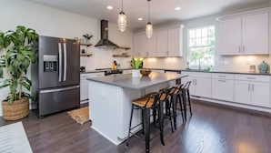 Una cocina moderna con armarios blancos, una isla central con taburetes, un refrigerador de acero inoxidable, iluminación colgante y un amplio ventanal que proporciona luz natural.