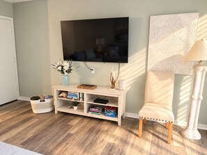 A living room with a flat-screen TV mounted on the wall, a wooden entertainment unit with books and decorative items, a cushioned armchair, and a patterned wall decoration.