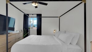 Ein modernes Schlafzimmer mit einem Himmelbett, Deckenventilator, Nachttisch mit Lampe und Blick ins Freie.