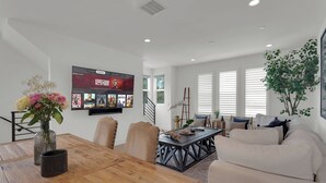 Ein modernes Wohnzimmer mit einem Holzesstisch, beigen Stühlen, einem weißen Sofa und einem an der Wand befestigten Flachbildfernseher, der einen Film zeigt.