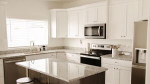 Cocina moderna con armarios blancos, una isla grande con taburetes, electrodomésticos de acero inoxidable y una ventana con persianas.
