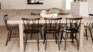 Un área de comedor con una mesa de madera y sillas negras, un sofá, un televisor montado en la pared y un mueble con objetos decorativos.