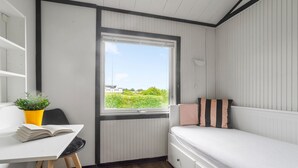 Une pièce de taille modeste, comprenant un lit, un bureau agrémenté d’une plante et une fenêtre donnant sur un paysage verdoyant et des bâtiments.
