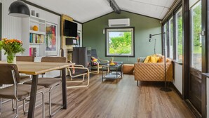 Un salon moderne avec un plancher en bois, un coin repas comprenant une table et des chaises, un canapé et une grande fenêtre donnant sur un paysage verdoyant.