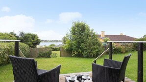 Une terrasse dotée de chaises et d’une table en osier, donnant sur un espace verdoyant, avec vue sur une étendue d’eau et un bâtiment situé au loin.