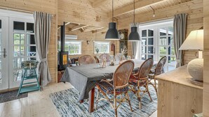 L’intérieur d’un chalet en bois chaleureux et accueillant comprend un coin repas avec des chaises en osier, une table recouverte d’une nappe grise et une grande fenêtre agrémentée de rideaux.