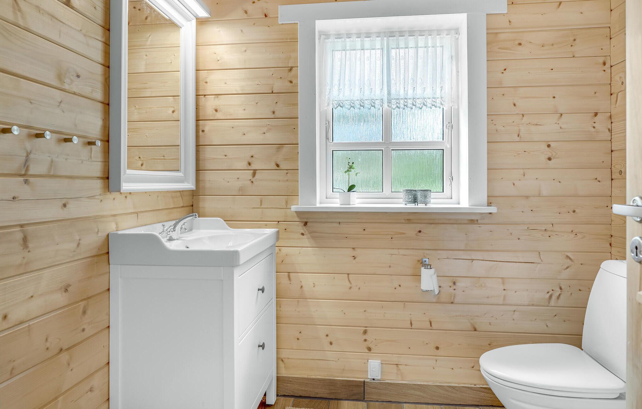 A bathroom with wooden walls, a white sink, a window with lace curtains, and a toilet.