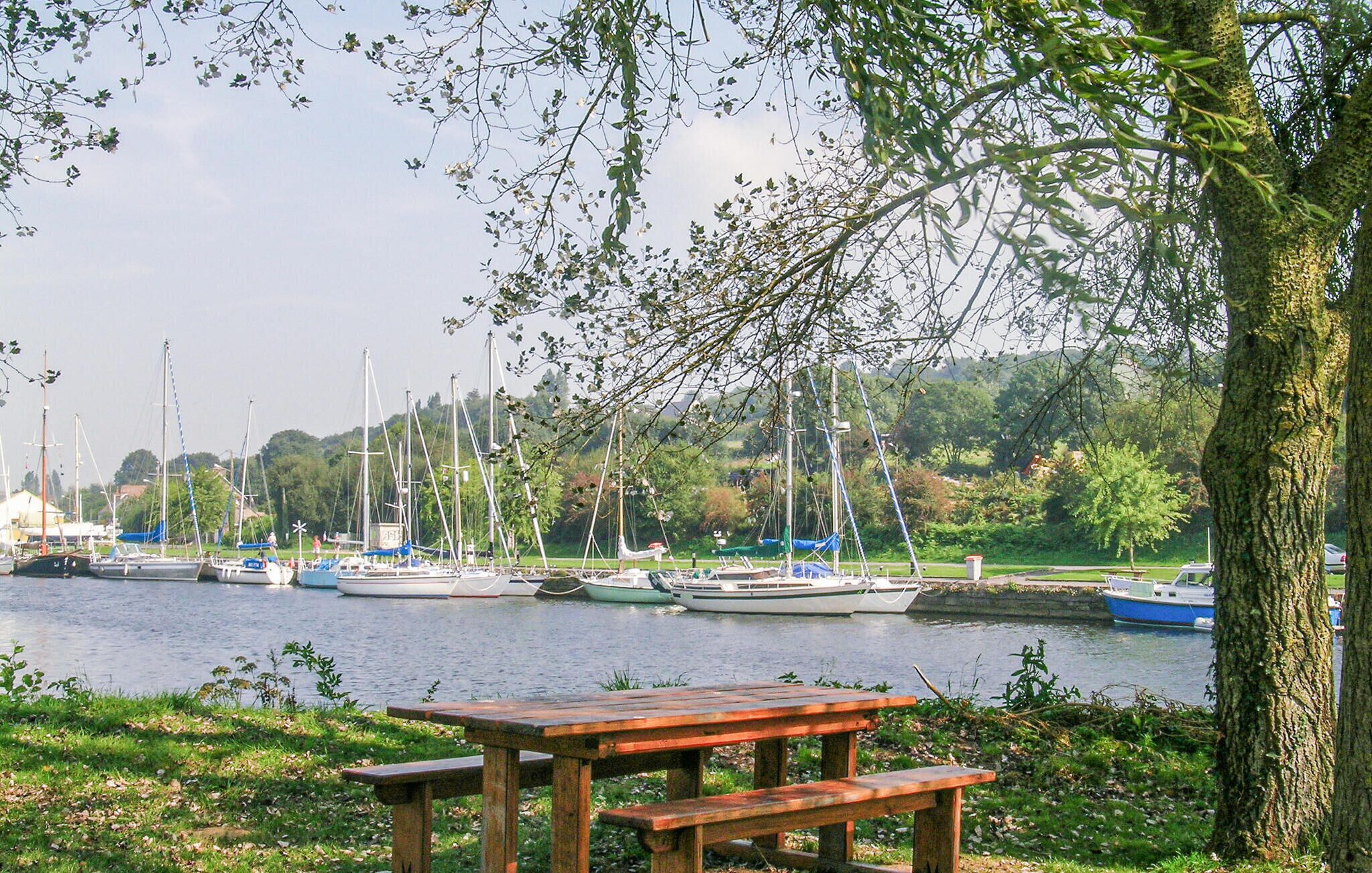 Un espace de pique-nique au bord de la rivière, avec un banc en bois et une vue sur les voiliers qui naviguent sur l’eau.
