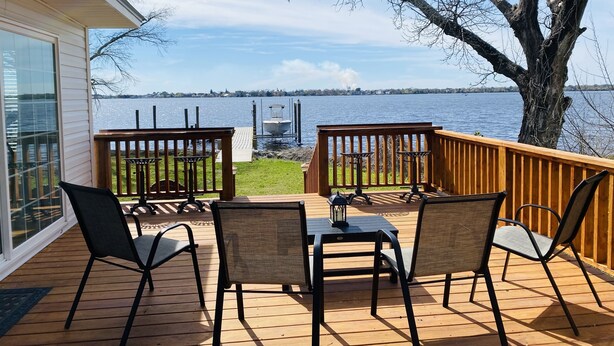 Une terrasse en bois avec une table et des chaises, donnant sur un lac où se trouvent un quai et des bateaux.