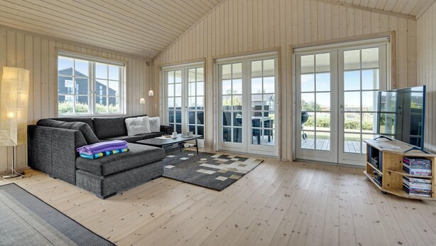 Un salón espacioso con un sofá gris, una mesa de centro y un mueble para la televisión con libros. Amplias ventanas y puertas correderas ofrecen abundante luz natural y vistas.