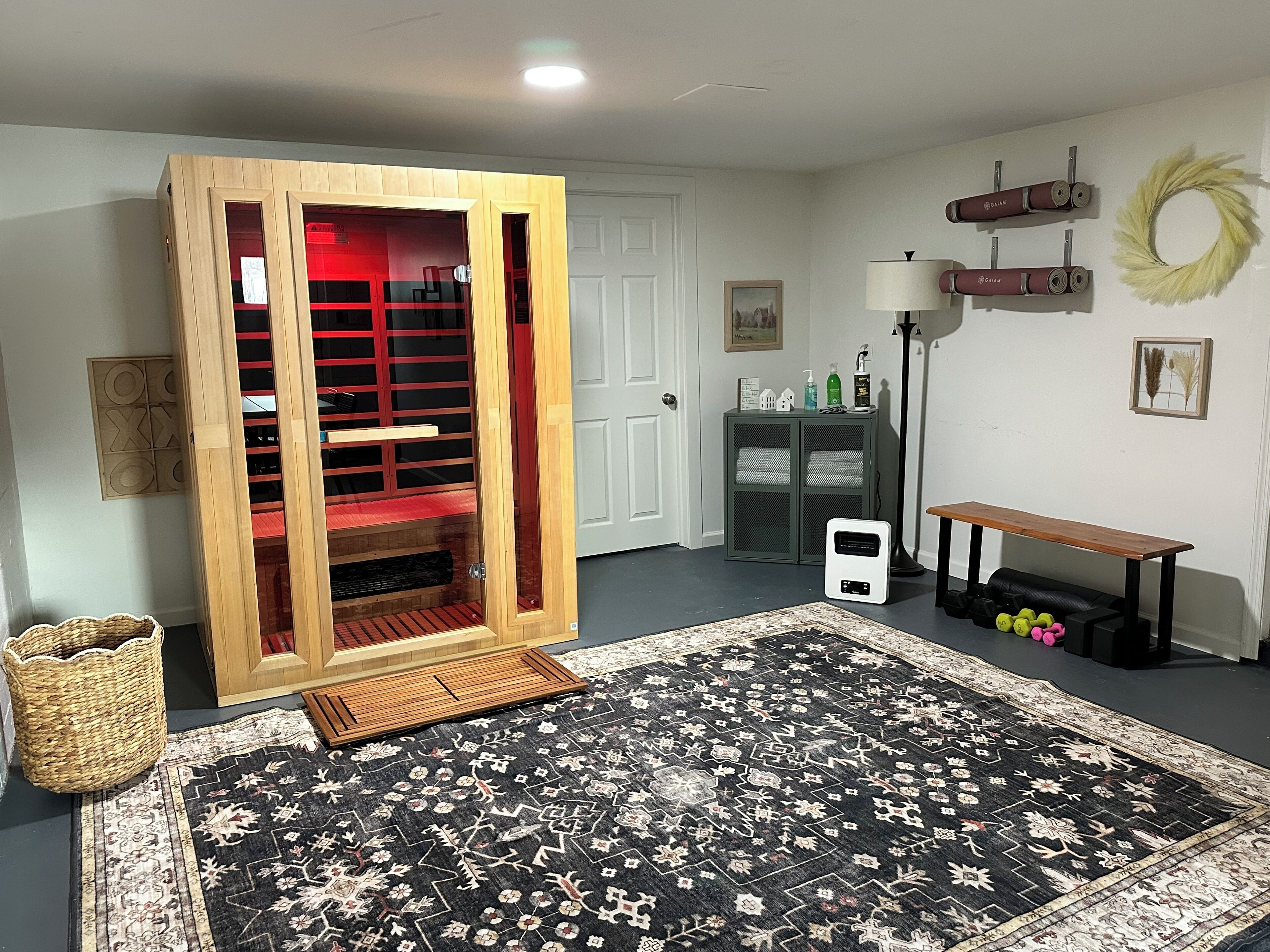 Una habitación con un gran armario de madera, una alfombra con estampado blanco y negro, un pequeño calentador blanco, un banco, una lámpara y un estante de pared con adornos.
