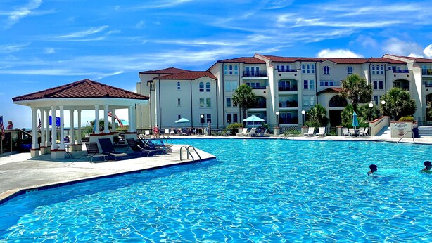 Una piscina con sillas de descanso y un pabellón, rodeada de edificios de varios pisos con balcones y un cielo azul despejado.