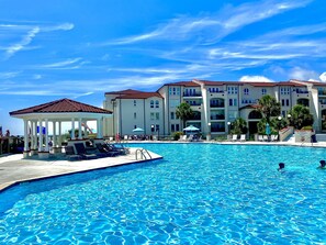 A swimming pool with lounge chairs and a gazebo, surrounded by multi-story buildings with balconies and a clear blue sky. - Oceanfront; 2 King Suites; Top Floor Corner Unit; Elevator (North Topsail Beach)