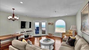 Una sala de estar con piso de madera, sofás color beige, una mesa de centro, un televisor y vista al océano a través de un ventanal grande.