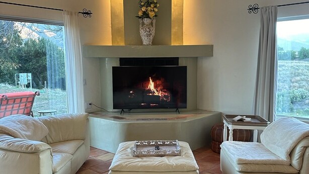 Un salon chaleureux avec une cheminée, deux canapés blancs, une table basse et un écran de télévision diffusant une image de feu.