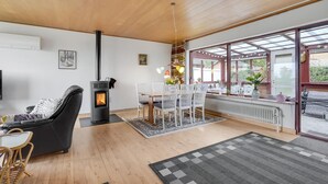 Een gezellige woonkamer met een houtkachel, een comfortabele bank, een eethoek met stoelen en een groot raam met uitzicht op de natuur.