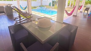 Un espacio al aire libre cubierto con una mesa de vidrio, sillas de mimbre, una piscina y hamacas.