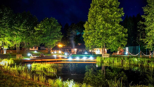A nighttime view of a lakeside area with a wooden dock, lounge chairs, and a building with outdoor lighting.