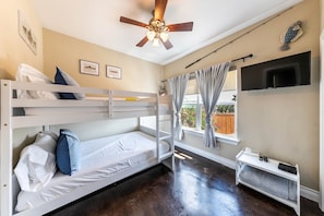 A room with bunk beds, a ceiling fan, a TV mounted on the wall, and a window with curtains.