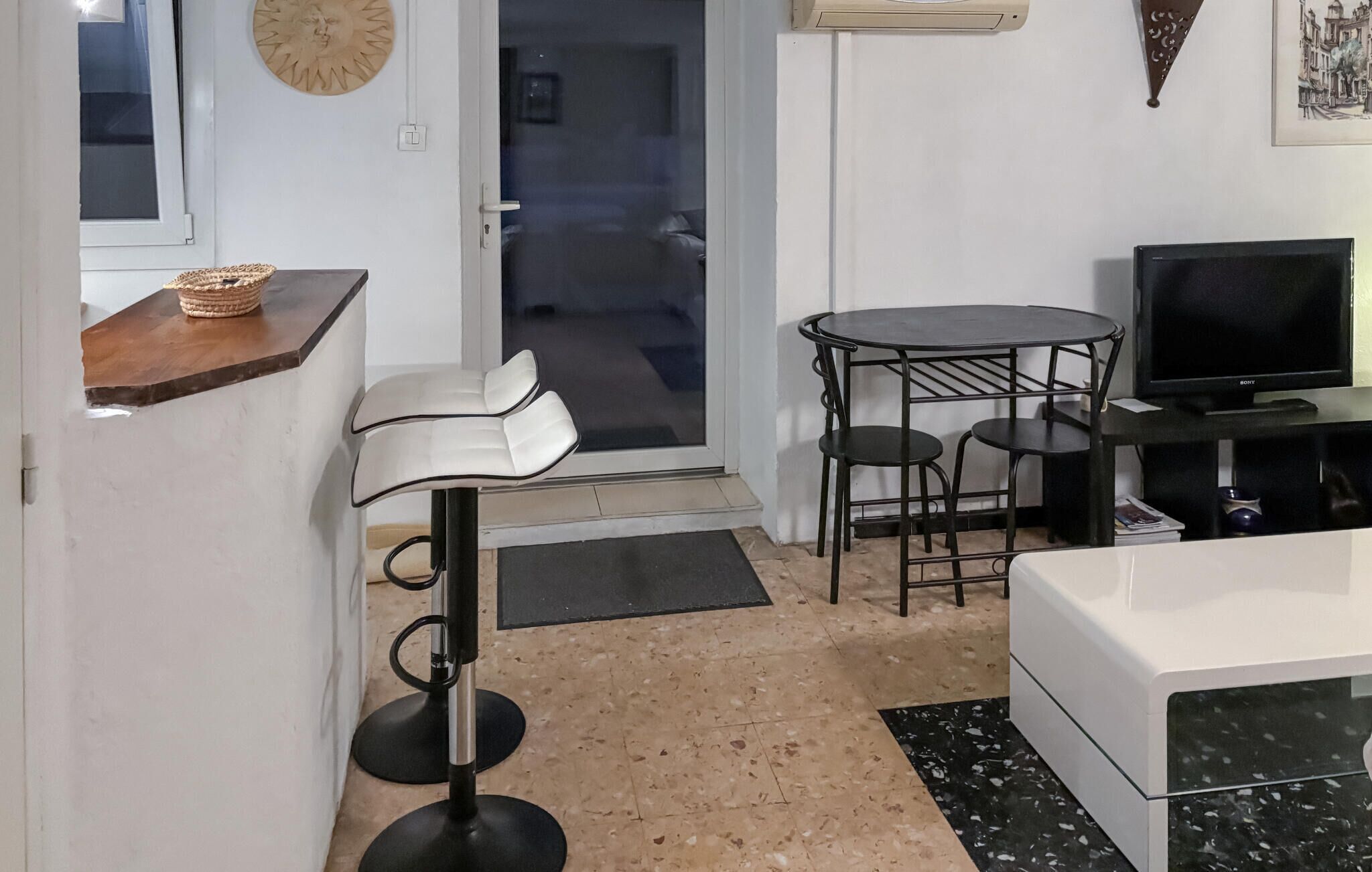 Een compact woongedeelte met een kitchenette, een eethoek met een ronde tafel en stoelen, een bank, een televisie en een decoratief wandobject.