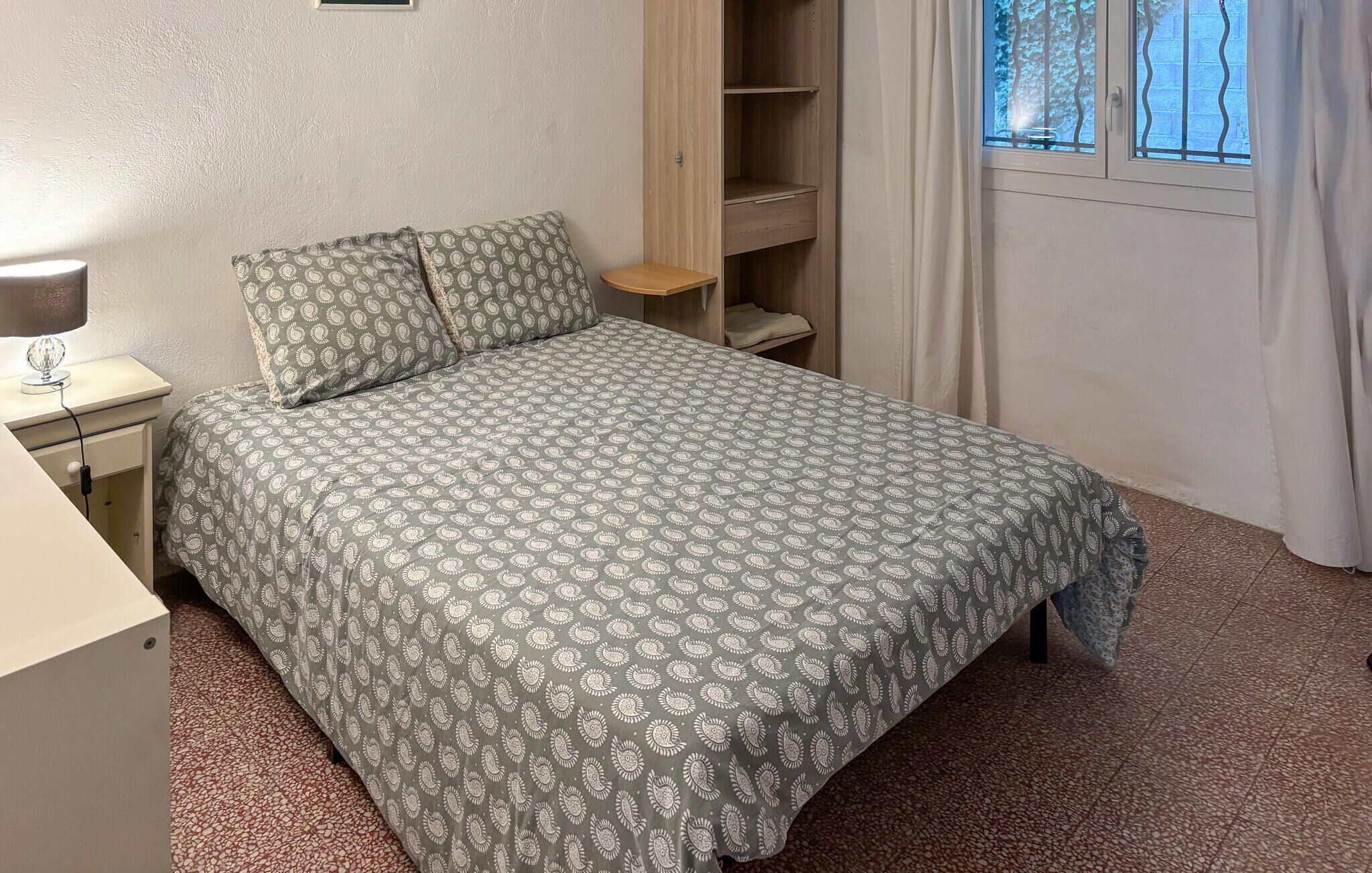 A bedroom with a bed, a nightstand, a wooden shelf, and a window with curtains.