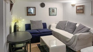 Una sala de estar con un sofá azul, un sofá blanco, una mesita y un armario blanco. Hay dos relojes en la pared y un cuadro enmarcado sobre el sofá blanco.