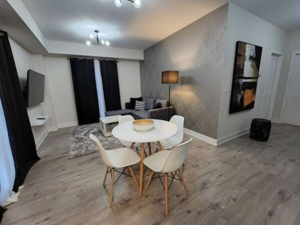 A modern living room with a dining area, featuring a round table with chairs, a sofa, a flat-screen TV, and a painting on the wall.