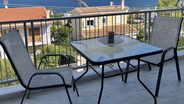 Een balkon met een glazen tafel en twee stoelen, met uitzicht op de kust, gebouwen en de zee.
