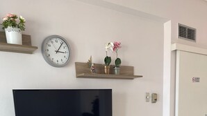 Een moderne woonkamer met een flatscreen televisie, een eettafel met stoelen, een klein bijzettafeltje en decoratieve elementen, waaronder een klok en potplanten.