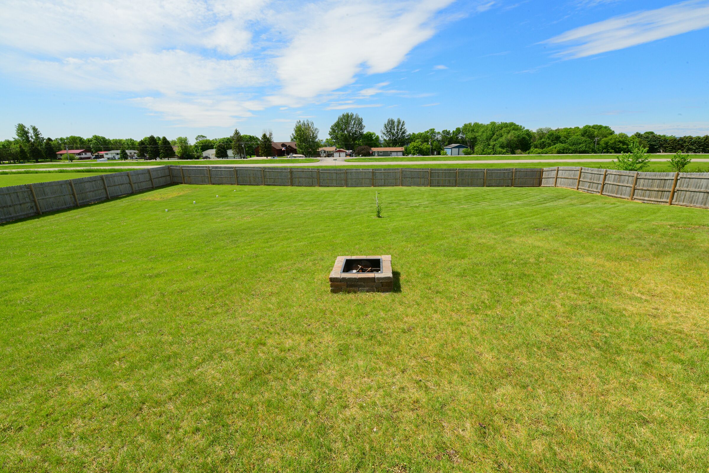 A grassy backyard with a fire pit, a wooden fence, and houses in the background.