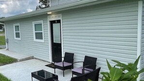 Un petit bâtiment avec une porte fermée, une terrasse en béton et du mobilier d’extérieur comprenant une table et des chaises.