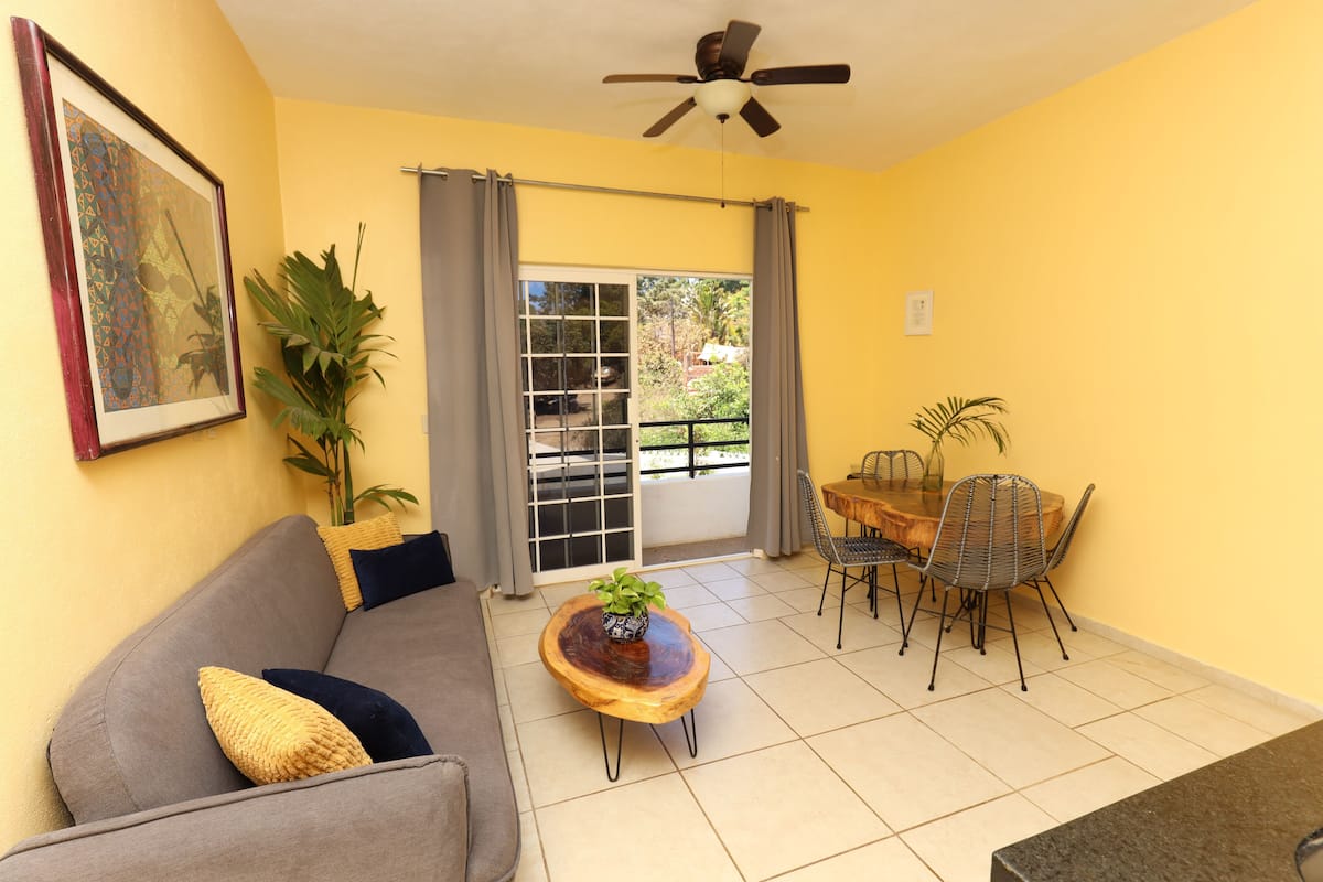 A living room with a sofa, a coffee table, a dining table with chairs, a ceiling fan, and a sliding glass door leading to an outdoor area.