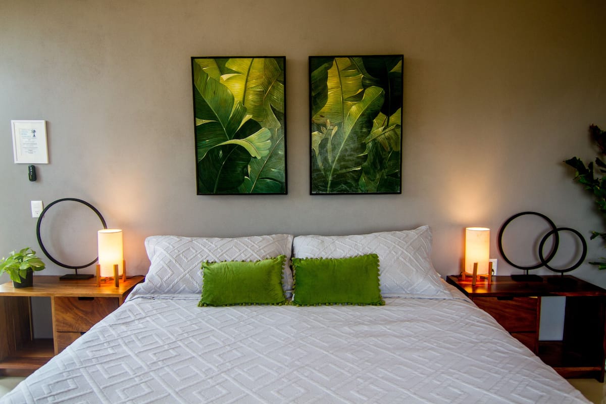 A neatly made bed with white bedding and two green pillows, flanked by bedside tables with lamps, and two framed botanical prints on the wall.