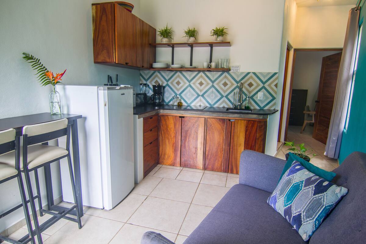A compact kitchen with a refrigerator, a coffee maker, and a sink. There is a wooden cabinet above the counter and a small shelf with plants. A purple sofa with a patterned pillow is in the foreground.