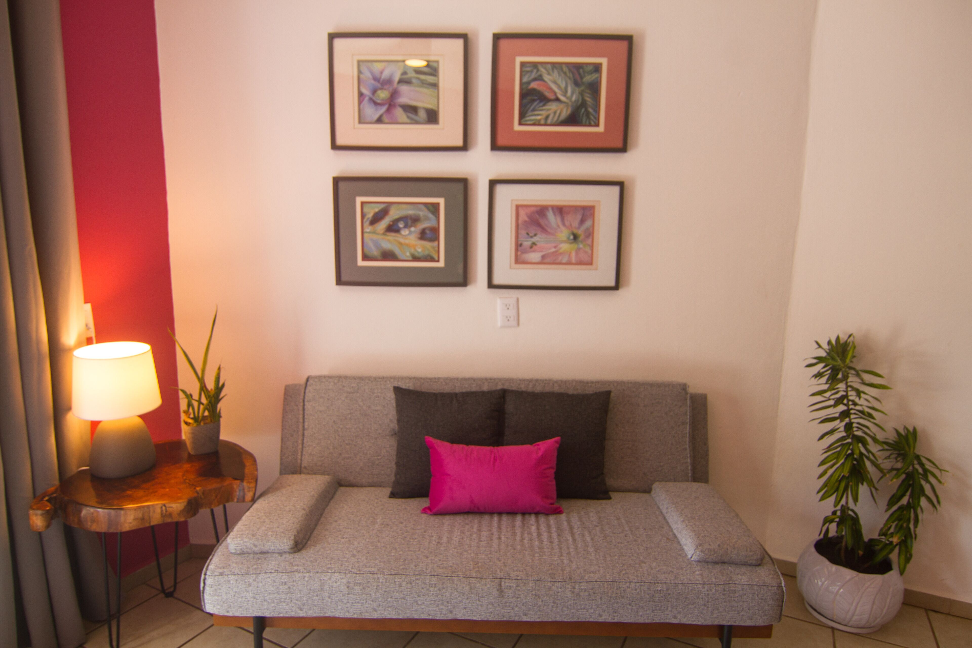 A modern living room with a grey sofa, a pink pillow, a wooden side table with a lamp, and four framed artworks on the wall.