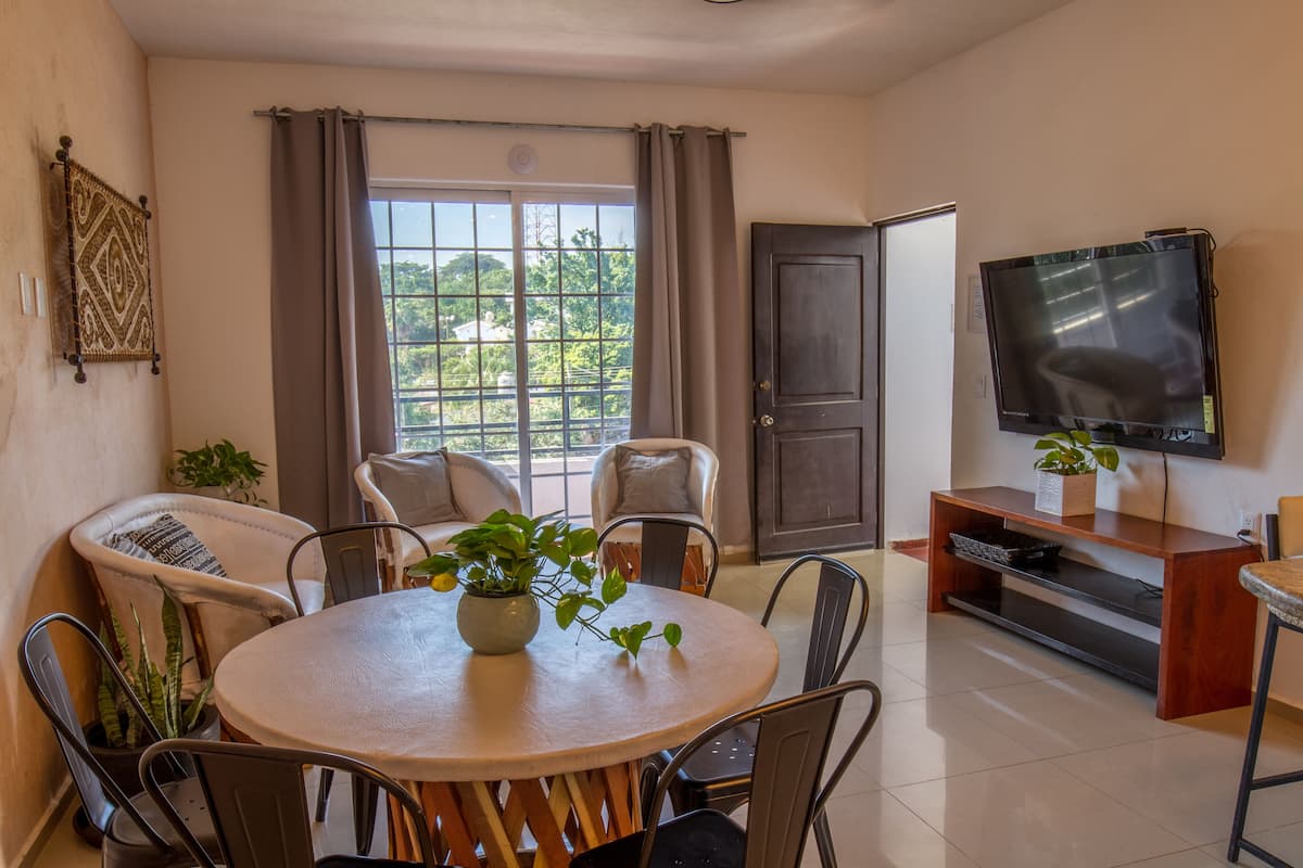 A dining area with a round table, chairs, a TV, and a view of greenery outside.